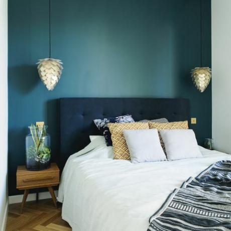 Interior de quarto elegante com pequena mesa de cabeceira de madeira, jardim em uma jarra, roupa de cama branca, pilhas de cores e cobertor. Espaço com paredes azuis e parquet de madeira marrom. Lâmpada de design.