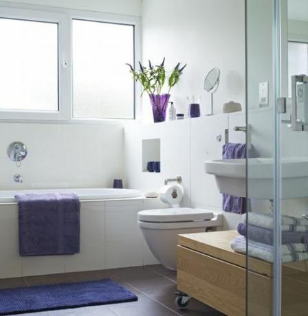 Banheiro bem iluminado com uma toalha roxa ao lado da banheira e toalhas dobradas perto do chuveiro