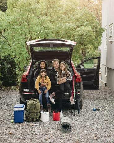 pais sorridentes sentados com as filhas no porta-malas de um carro elétrico