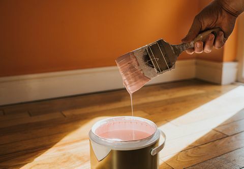 mão segura um pincel grosso e o remove, após mergulhá-lo na tinta, ele cria uma gota de tinta que respinga de volta ao pote, a parede atrás é pintada de laranja e a cor da lata é rosa, o que se choca conceitualmente com o espaço para cópia de