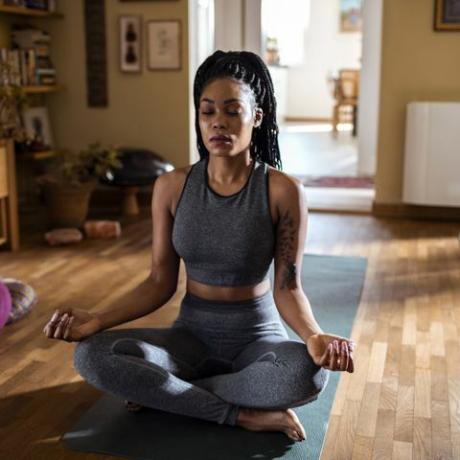 mulher negra sentada em uma sala ensolarada meditando na posição de lótus