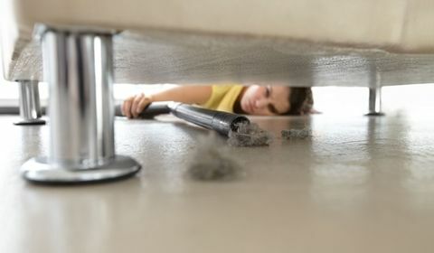 Mulher alcançando debaixo de um sofá para obter um pouco de poeira