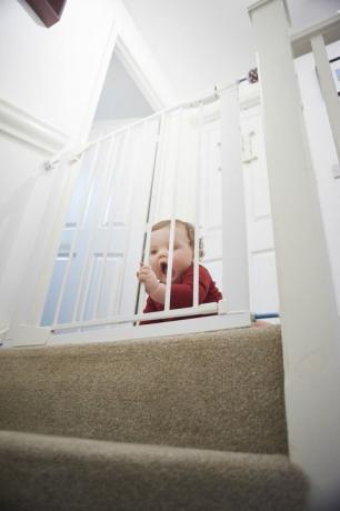 Portão de segurança do bebê nas escadas: Menino no patamar de sua casa. Ele está atrás de um portão de segurança para bebês que o impede do perigo da escada.