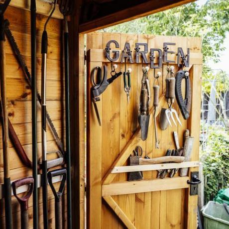 vista interna com porta aberta de galpão organizado cheio de ancinho, pás, aparador, serra e outras ferramentas manuais necessárias para a manutenção do jardim bem cuidado